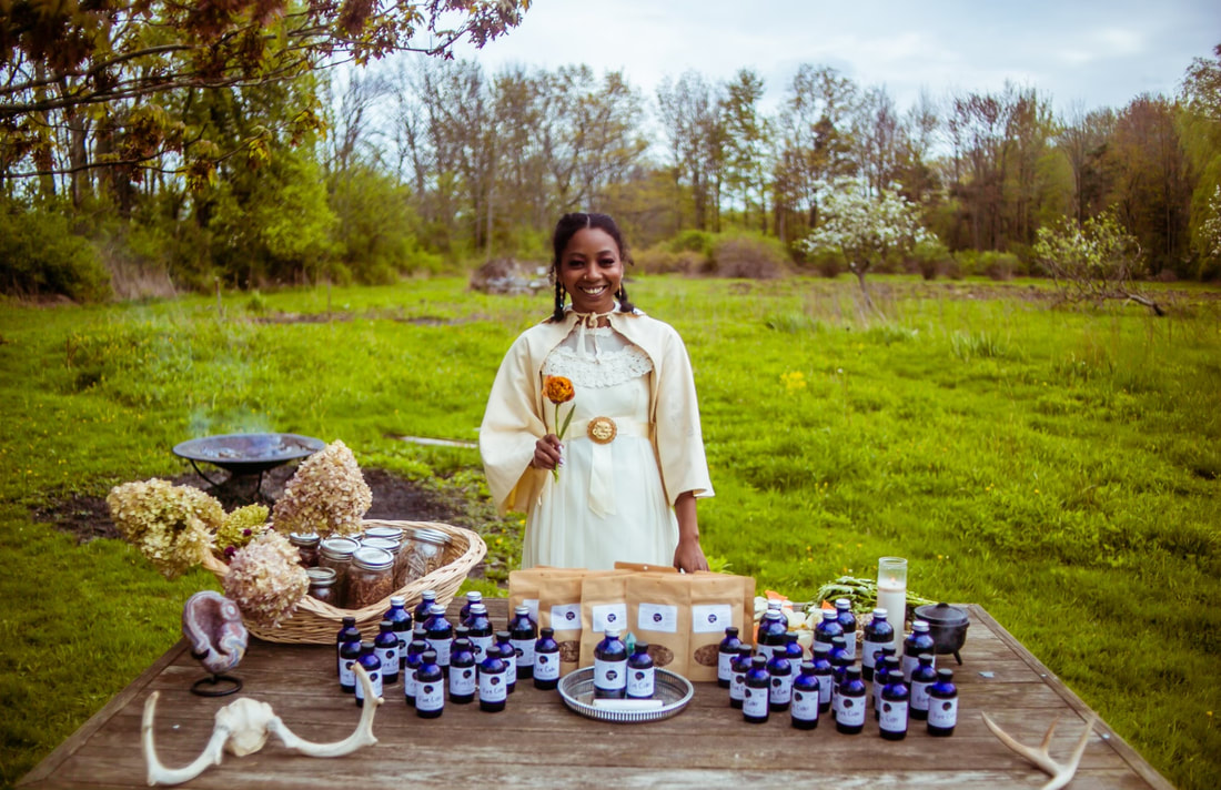 Nnenna stands in a green field of grass and trees, wearing a white dress, behind a wood table with a basket of jars of herbs, pouches of tea, blue bottles of elixers, and animal bones.