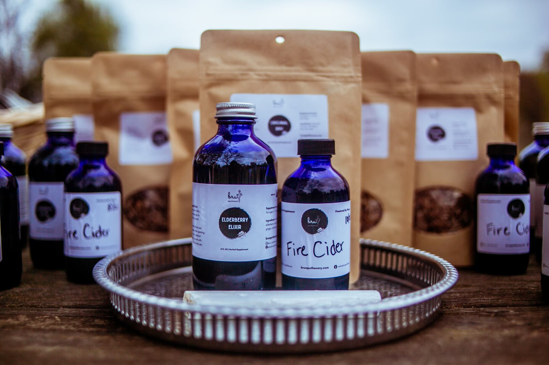 Dark blue bottles of Elderberry Elixer and Fire Cider are clustered in front of brown paper pouches of tea on a wood table.
