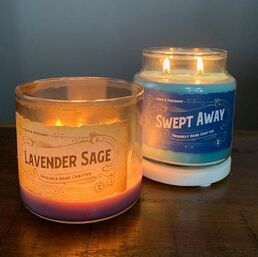 A mostly-burned Lavender Sage candle on the left and a blue lit Swept Away candle on the right, on a wood table with a grey wall behind.