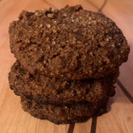 A stack of three dark brown cookies with grains of coarse sugar on top, placed on a light wood cutting board. 