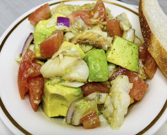 A salad of flaked white salted cod fish, green avocado pieces, red tomato pieces, purple red onion pieces, and a slice of bread. 