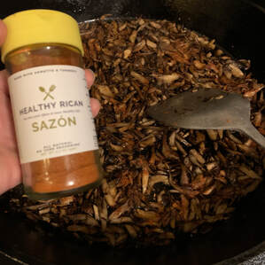 A hand holding a jar of sazon spice blend over a black cast iron pan full of sauteed mushrooms.
