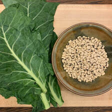 A, earth-toned pottery bowl holds dry black-eyed peas next to large dark green collard leaves on a wooden cutting board. 