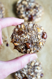 An index finger and thumb holding a textured oatmeal chia cookie with brown chocolate chips above a baking sheet and more cookies. 