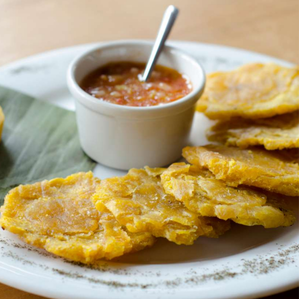 A white plate with several yellow tostones (fried plantains) with a small white dish of red sauce.s